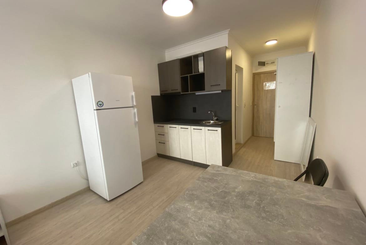 Современная студия с кухней, холодильником и обеденным столом, светлые тона, удобное планирование пространства.