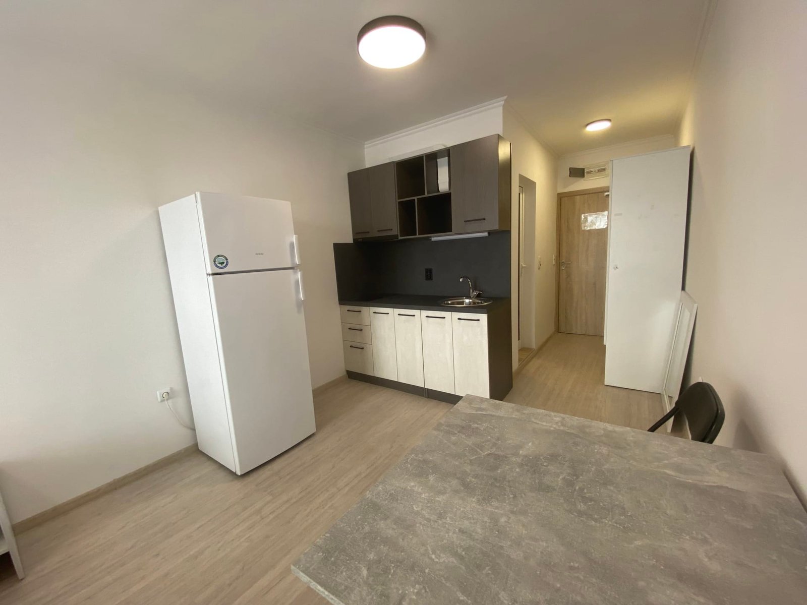 Современная студия с кухней, холодильником и обеденным столом, светлые тона, удобное планирование пространства.