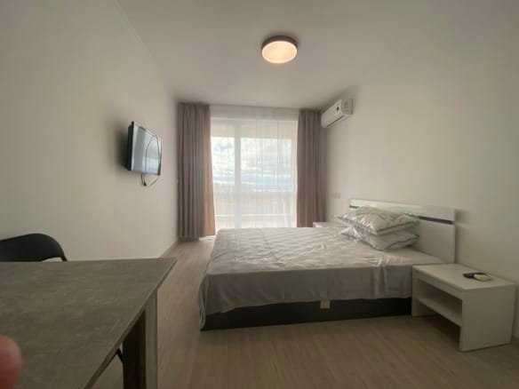 Современная студия с кроватью, телевизором на стене и окном, оформленная в светлых тонах.