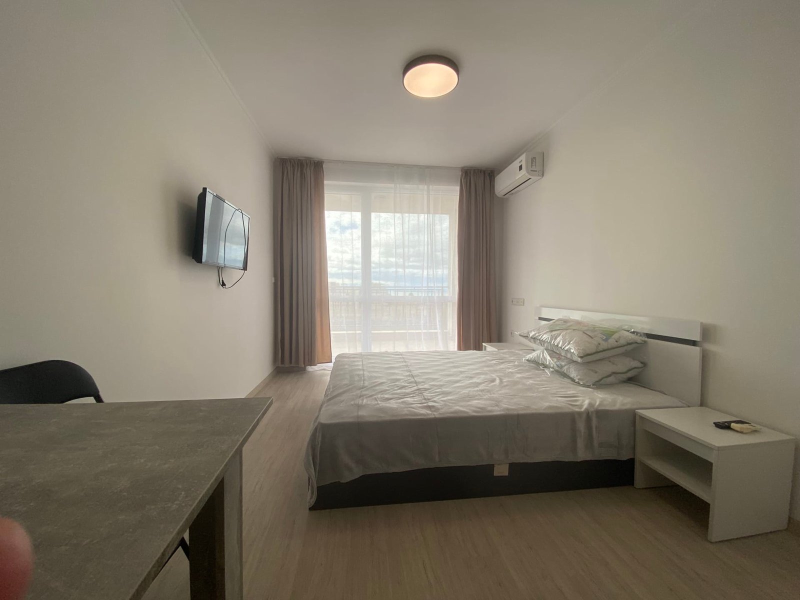 Современная студия с кроватью, телевизором на стене и окном, оформленная в светлых тонах.