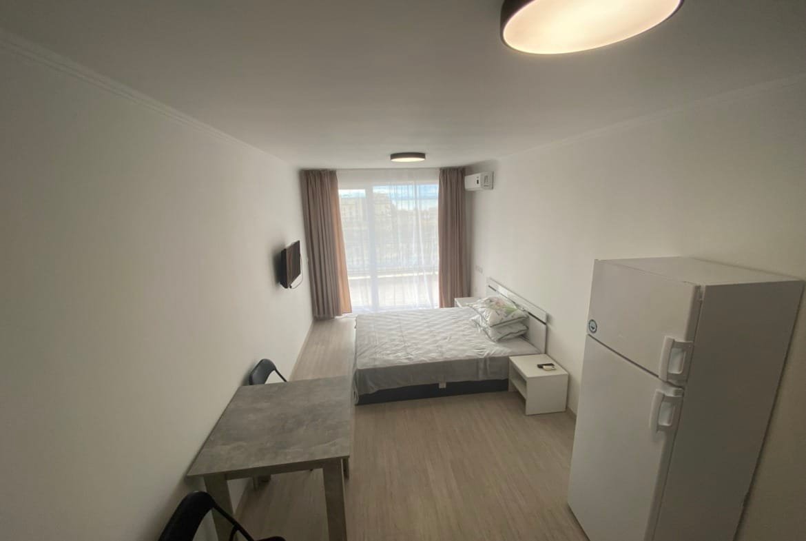 Современная студия с двуспальной кроватью, столом, стульем, холодильником и окном, пропускающим естественный свет.