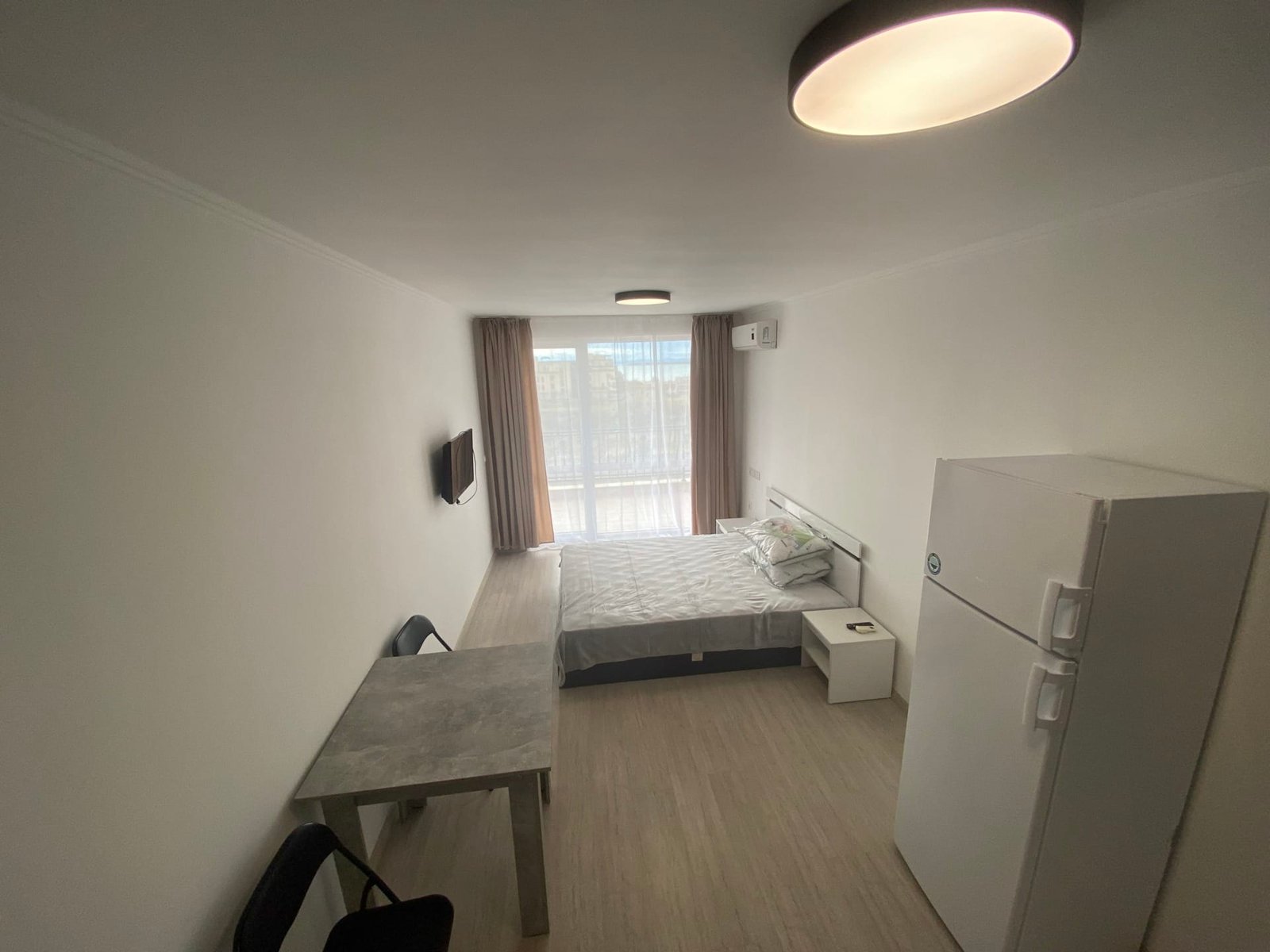 Современная студия с двуспальной кроватью, столом, стульем, холодильником и окном, пропускающим естественный свет.