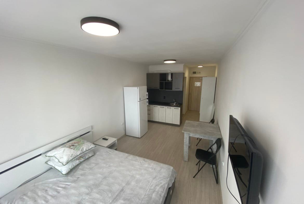 Современная студия с кухонной зоной, кроватью и обеденным столом, оформленная в светлых тонах.