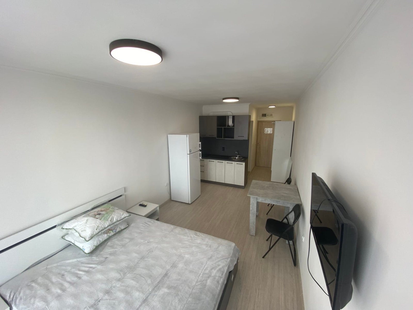 Современная студия с кухонной зоной, кроватью и обеденным столом, оформленная в светлых тонах.