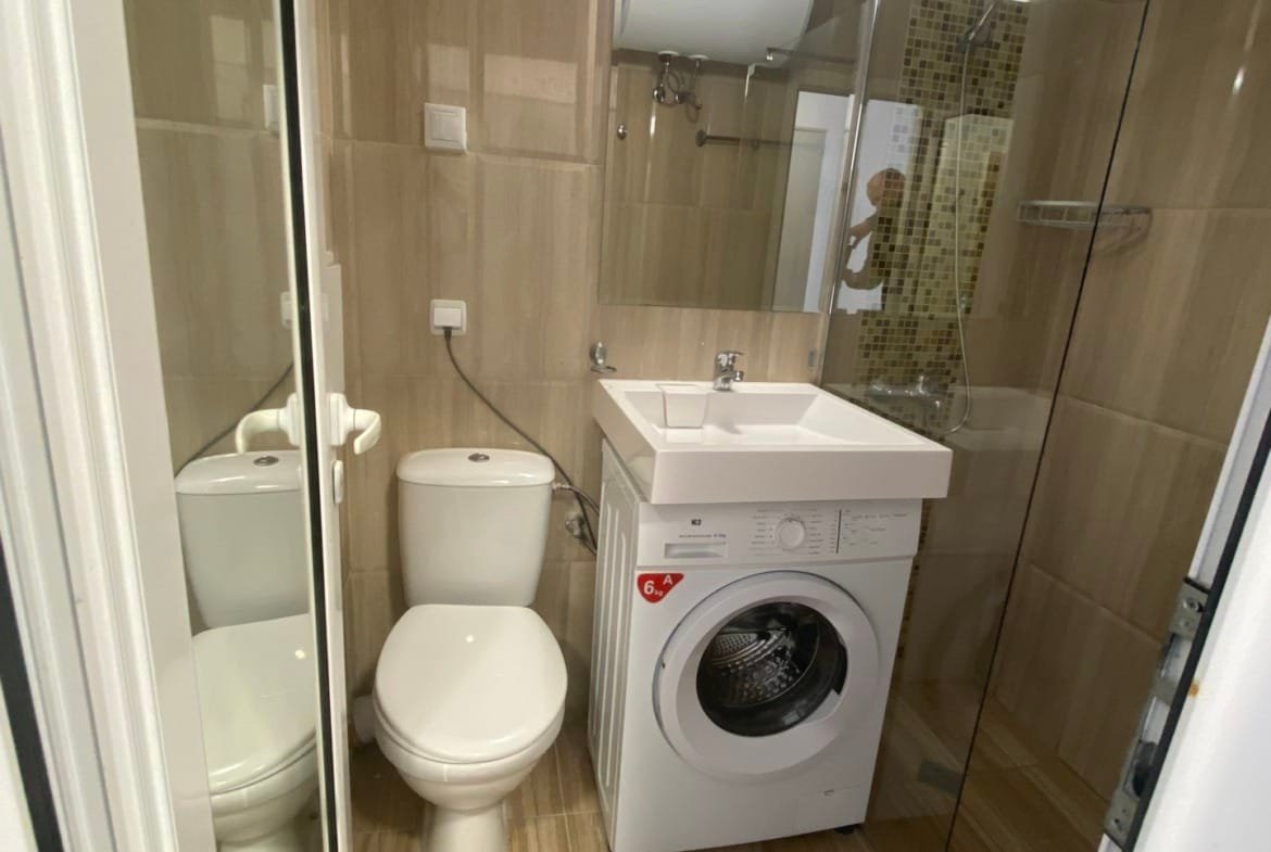 Современная ванная комната в студии с душевой кабиной, унитазом и стиральной машиной. Светлые плитки, аккуратный дизайн.