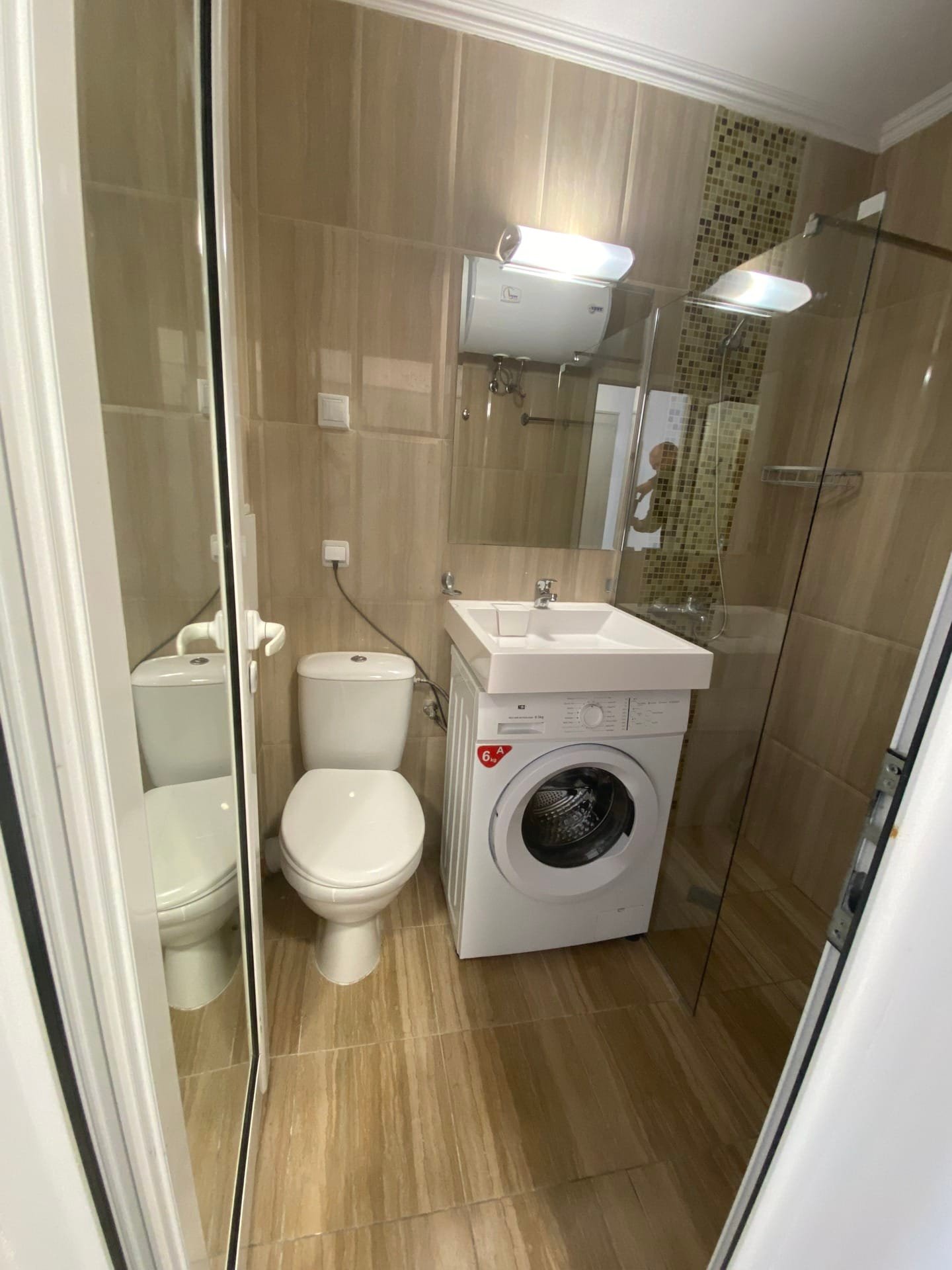 Современная ванная комната в студии с душевой кабиной, унитазом и стиральной машиной. Светлые плитки, аккуратный дизайн.