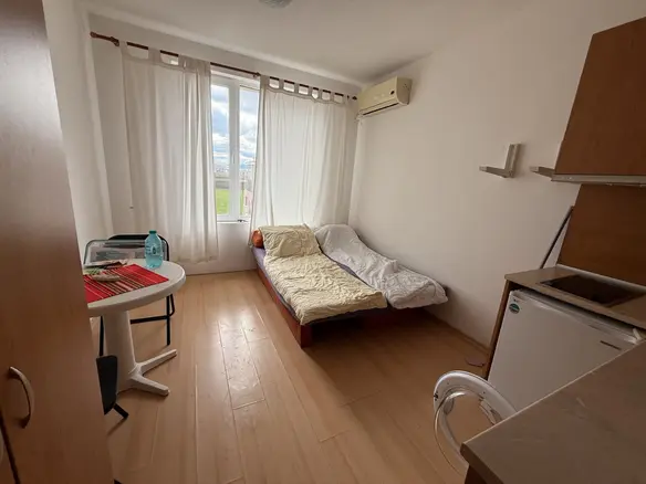 Уютная студия с кроватью, обеденным столом и кухонным уголком, яркие окна создают светлую атмосферу.