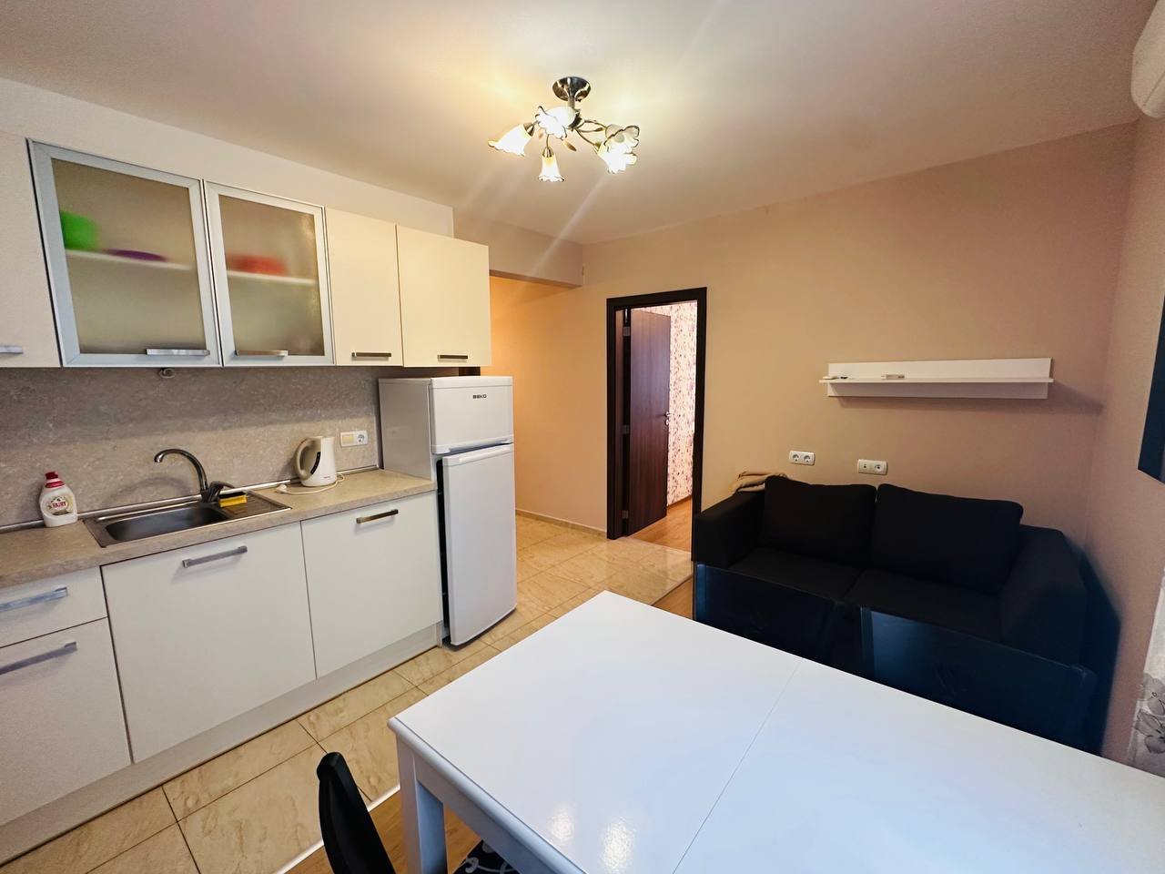 Современная кухня-студио с белыми шкаками, обеденным столом и черным диваном в апартаменте на побережье.