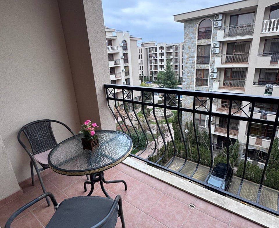 Балкон студии с пластиковыми стульями и стеклянным столом, цветы на столе, вид на жилой комплекс и зеленые насаждения.