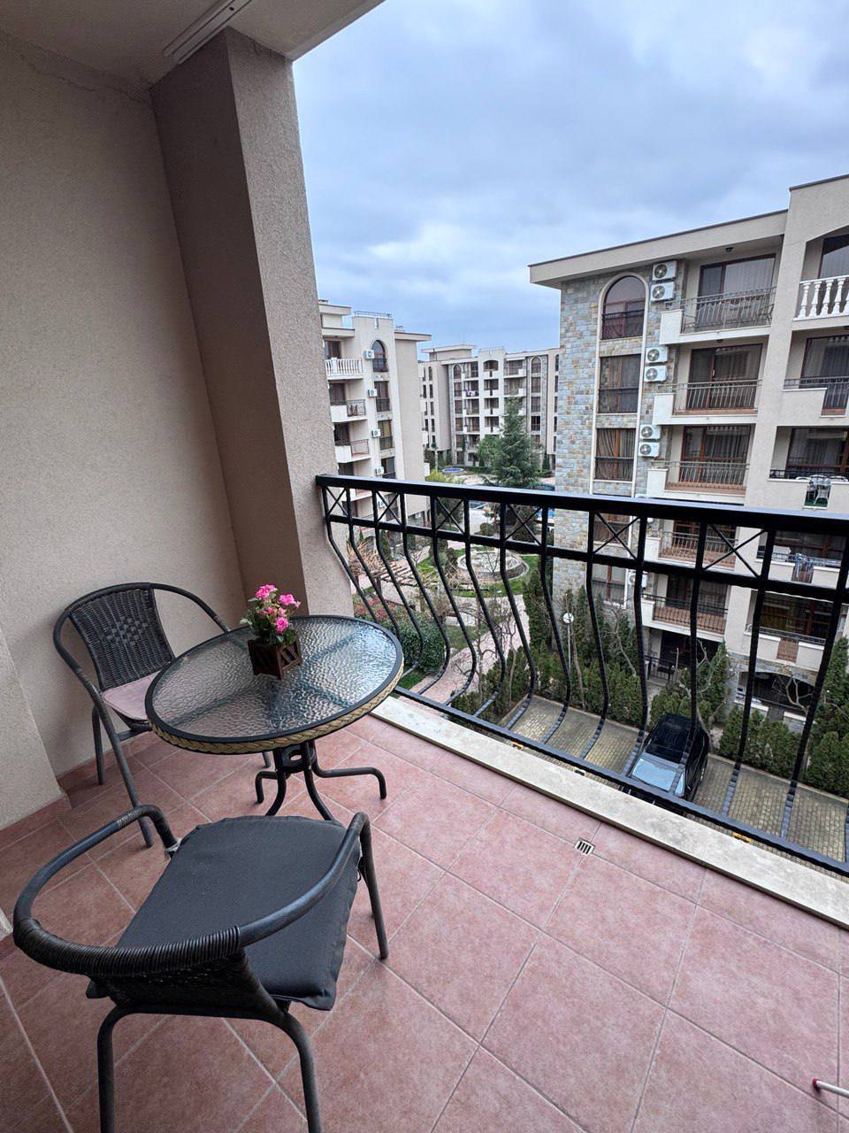 Балкон студии с пластиковыми стульями и стеклянным столом, цветы на столе, вид на жилой комплекс и зеленые насаждения.