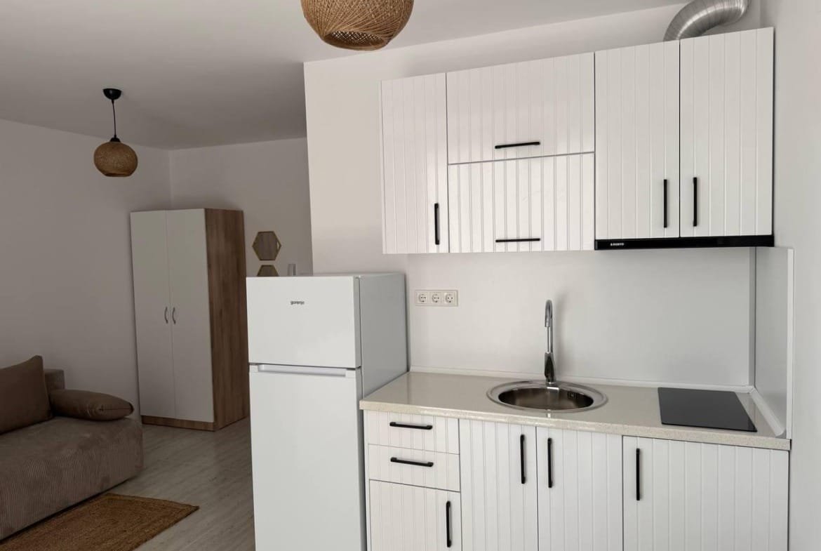 Современная кухня в студии с белыми шкафами, холодильником, мойкой и плитой, стилизованная в светлых тонах.