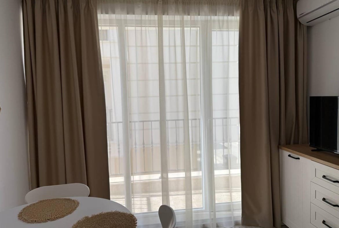 Современный интерьер студии с белыми и бежевыми шторами, круглым столом и телевизором на тёмном шкафу.