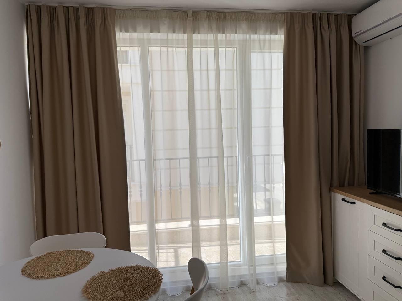 Современный интерьер студии с белыми и бежевыми шторами, круглым столом и телевизором на тёмном шкафу.