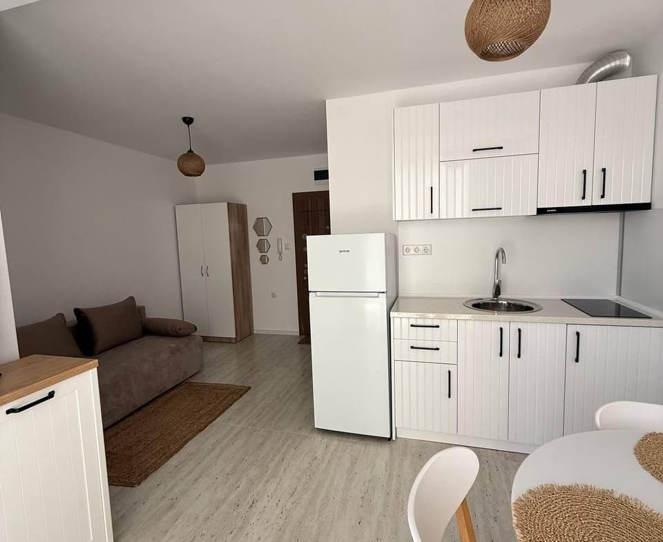 Современная студия с белыми кухонными шкафами, гладкой столешницей, диваном и обеденным столом на светлом фоне.