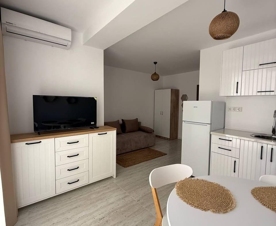 Современная студия с кухонным уголком, обеденным столом, диваном и телевизором в комплексе “Villa Grand”.