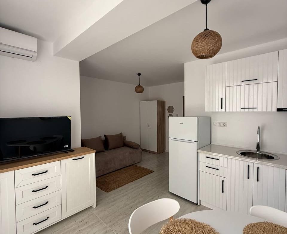 Современная студия с кухней, обеденным столом, диваном и тёплыми светильниками, оформленная в светлых тонах.