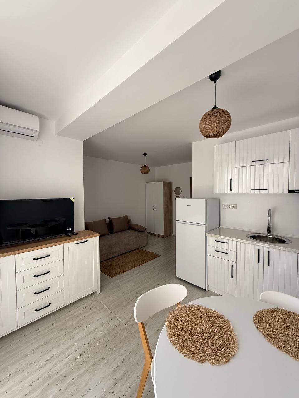 Современная студия с кухней, обеденным столом, диваном и тёплыми светильниками, оформленная в светлых тонах.