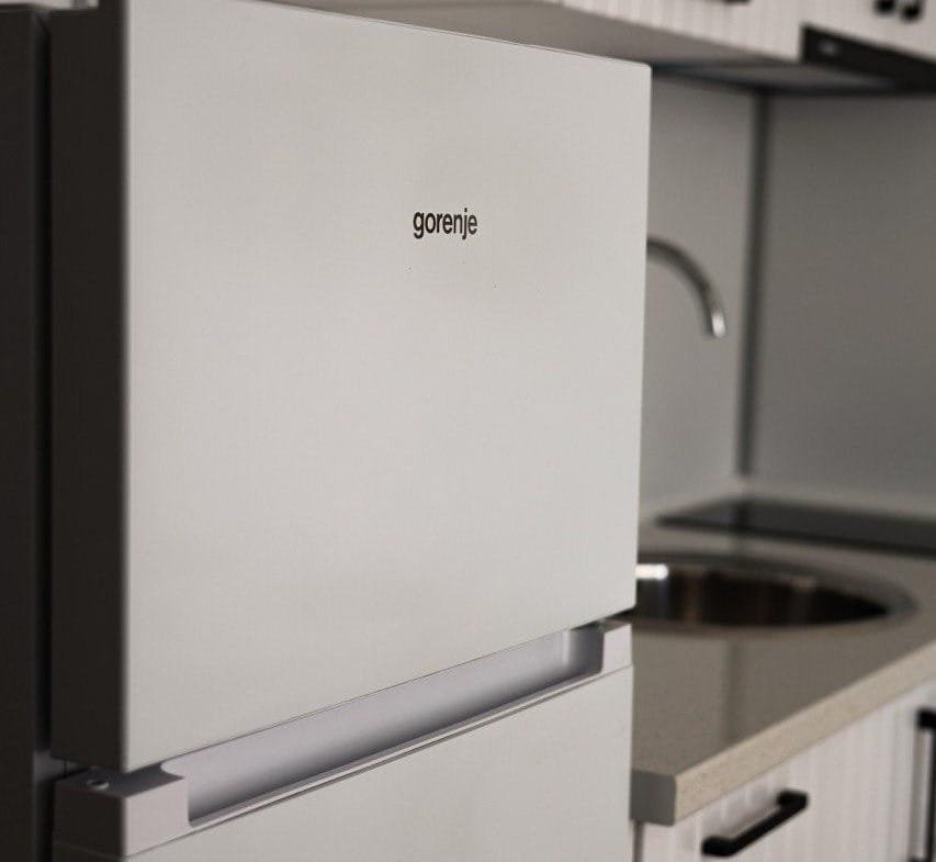 Белый холодильник Gorenje рядом с кухонной раковиной в студии Villa Grand, оформление в светлых тонах, современный стиль.