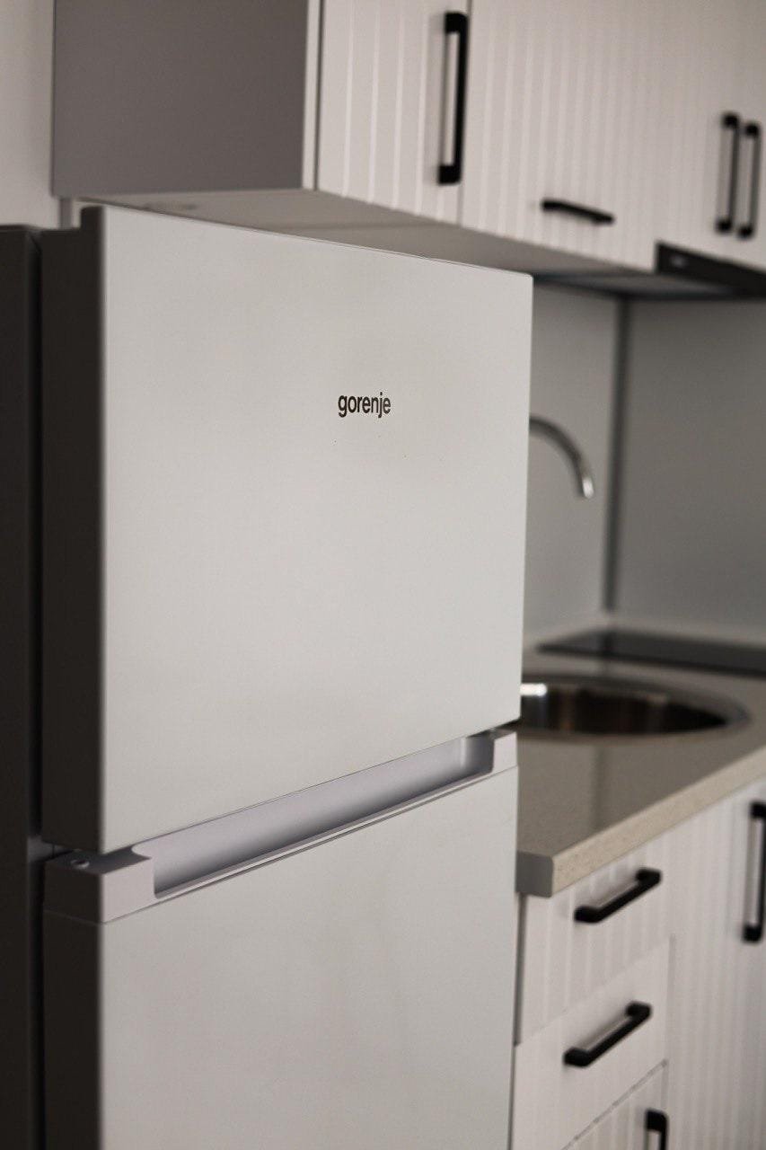 Белый холодильник Gorenje рядом с кухонной раковиной в студии Villa Grand, оформление в светлых тонах, современный стиль.