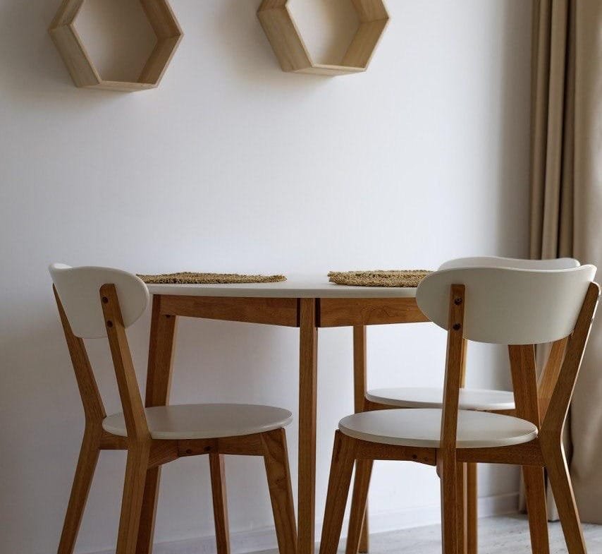 Деревянный круглый стол с двумя стульями в светлом интерьере, на стене висят hexagonal деревянные полки.