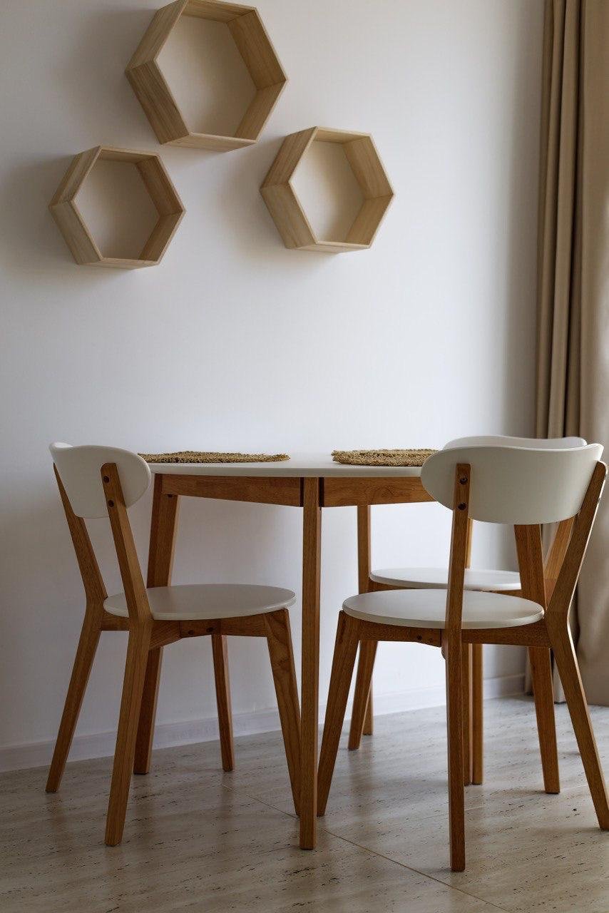 Деревянный круглый стол с двумя стульями в светлом интерьере, на стене висят hexagonal деревянные полки.