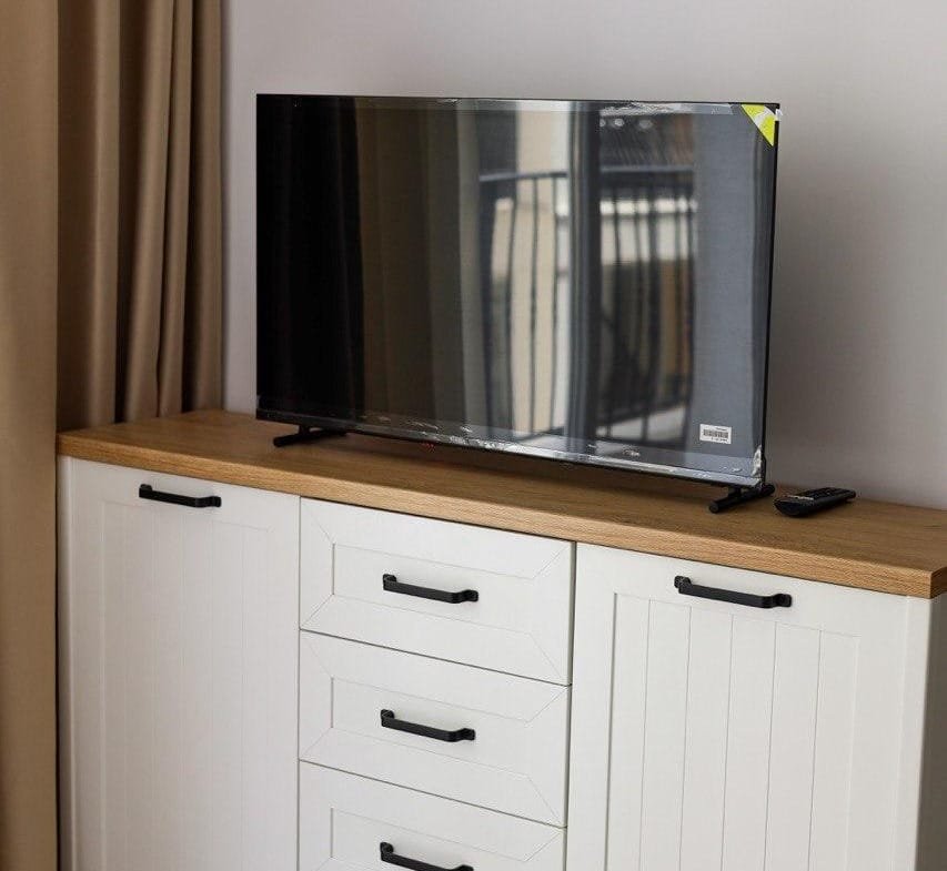 Телевизор на шкафу с деревянной столешницей, рядом пульт управления, светлые стены и занавески. Обстановка в квартире.