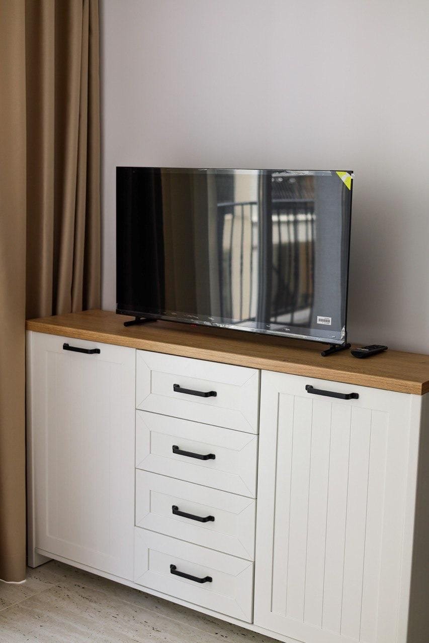 Телевизор на шкафу с деревянной столешницей, рядом пульт управления, светлые стены и занавески. Обстановка в квартире.