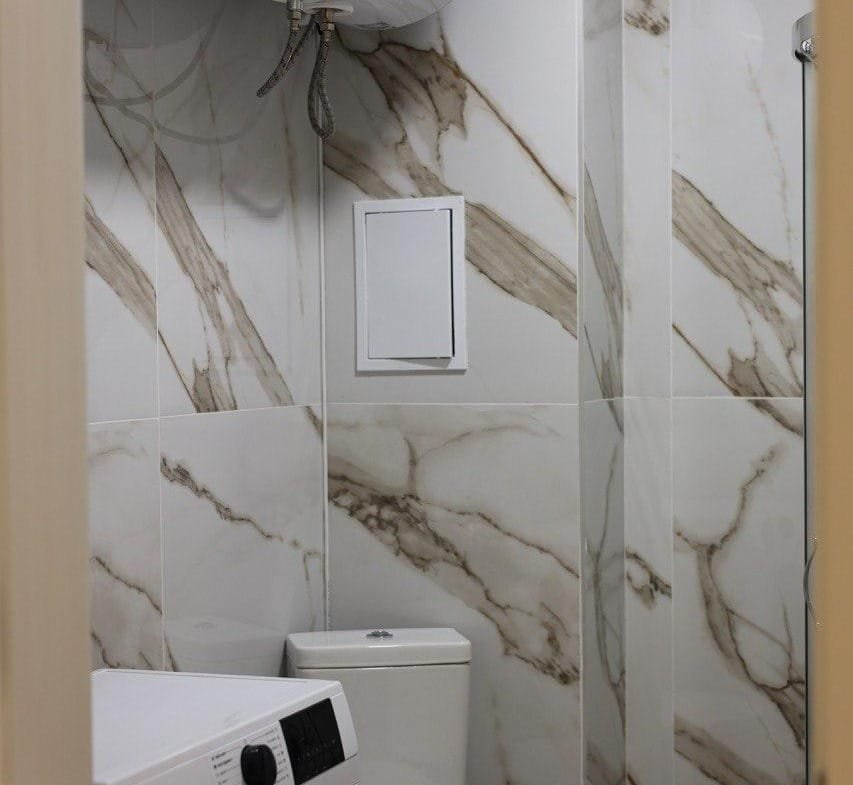 Современная ванная комната с белыми стенами, стиральной машиной и унитазом, отделанная мраморной плиткой.