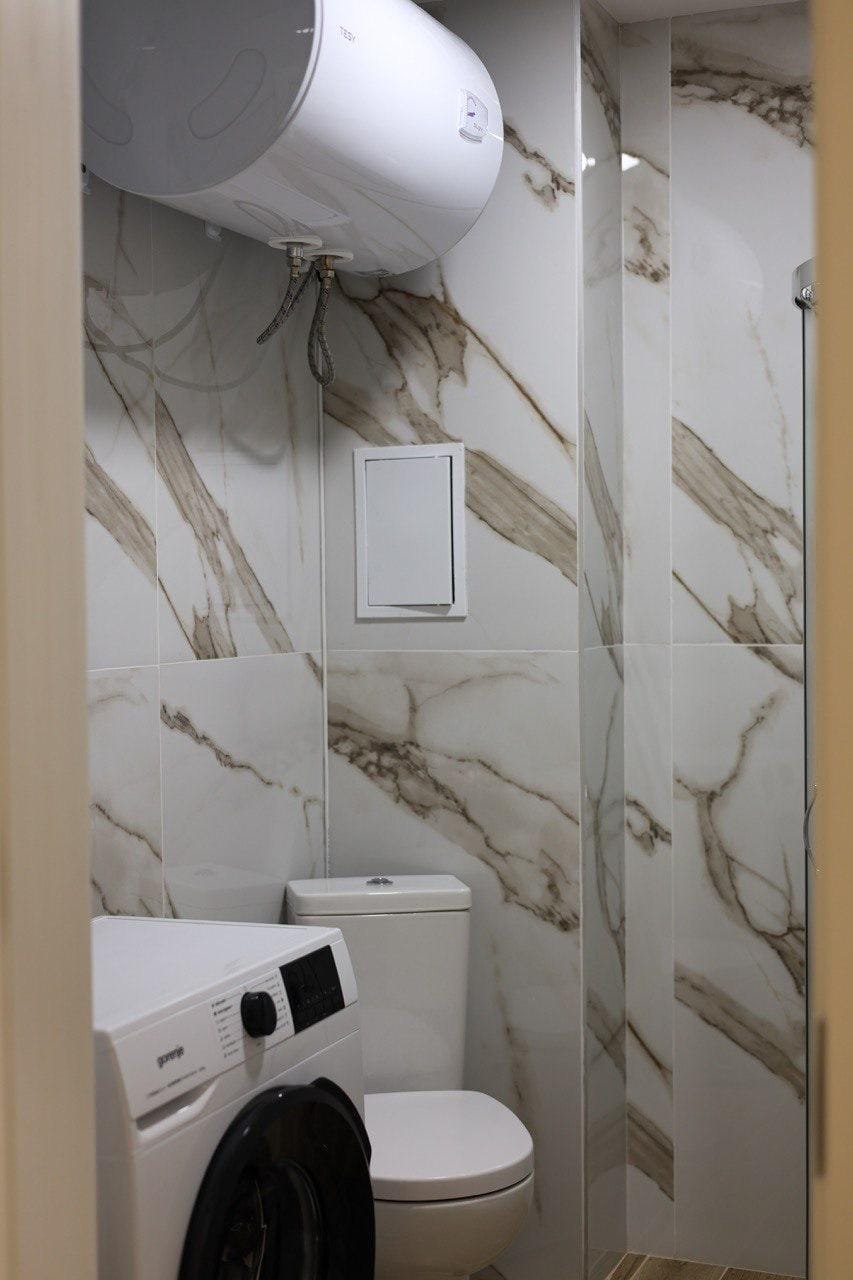 Современная ванная комната с белыми стенами, стиральной машиной и унитазом, отделанная мраморной плиткой.
