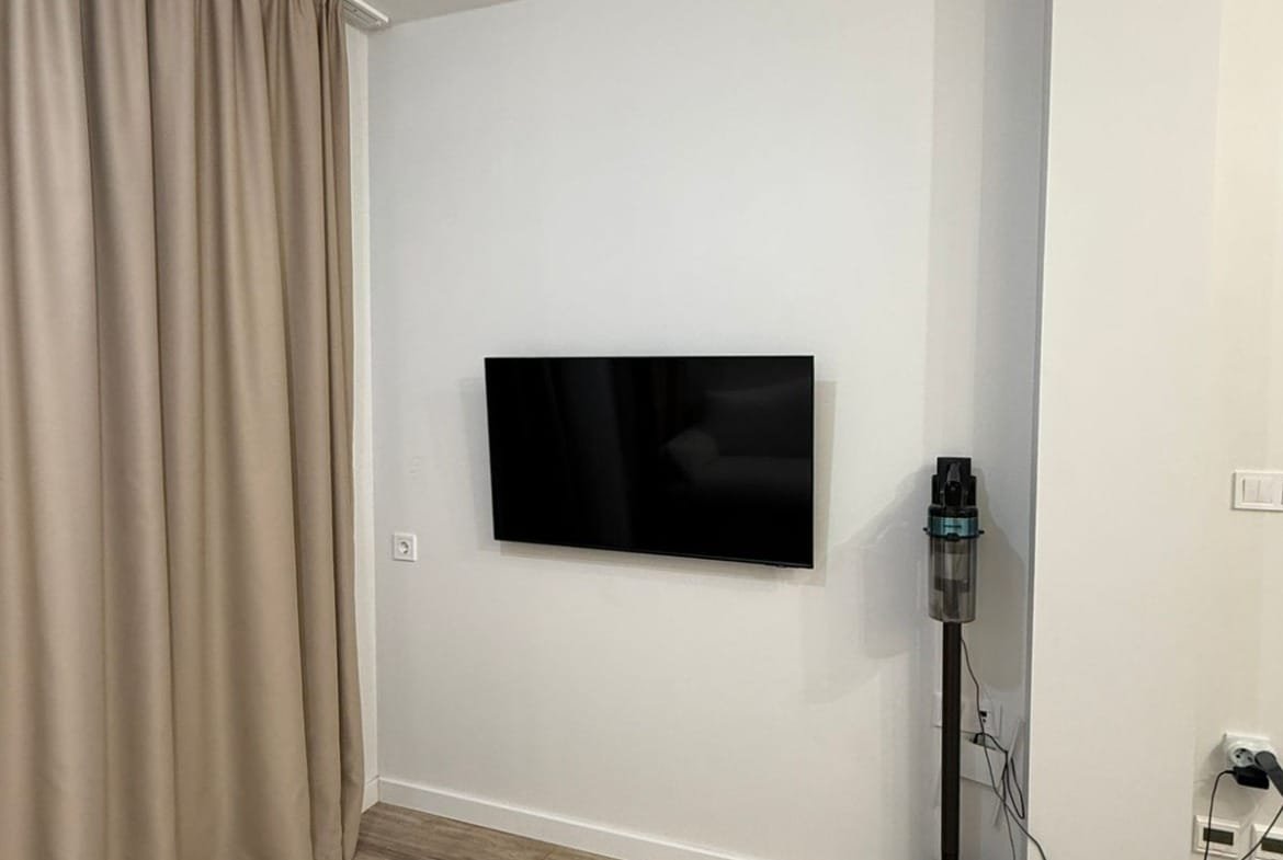 Светлая комната с настенным телевизором и аккуратным пылесосом, оформленная в современном стиле с элементами минимализма.