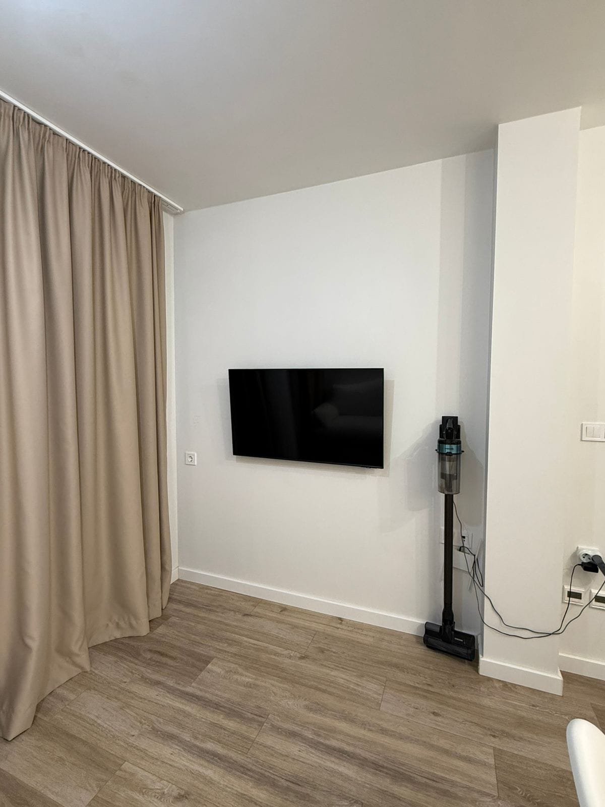 Светлая комната с настенным телевизором и аккуратным пылесосом, оформленная в современном стиле с элементами минимализма.