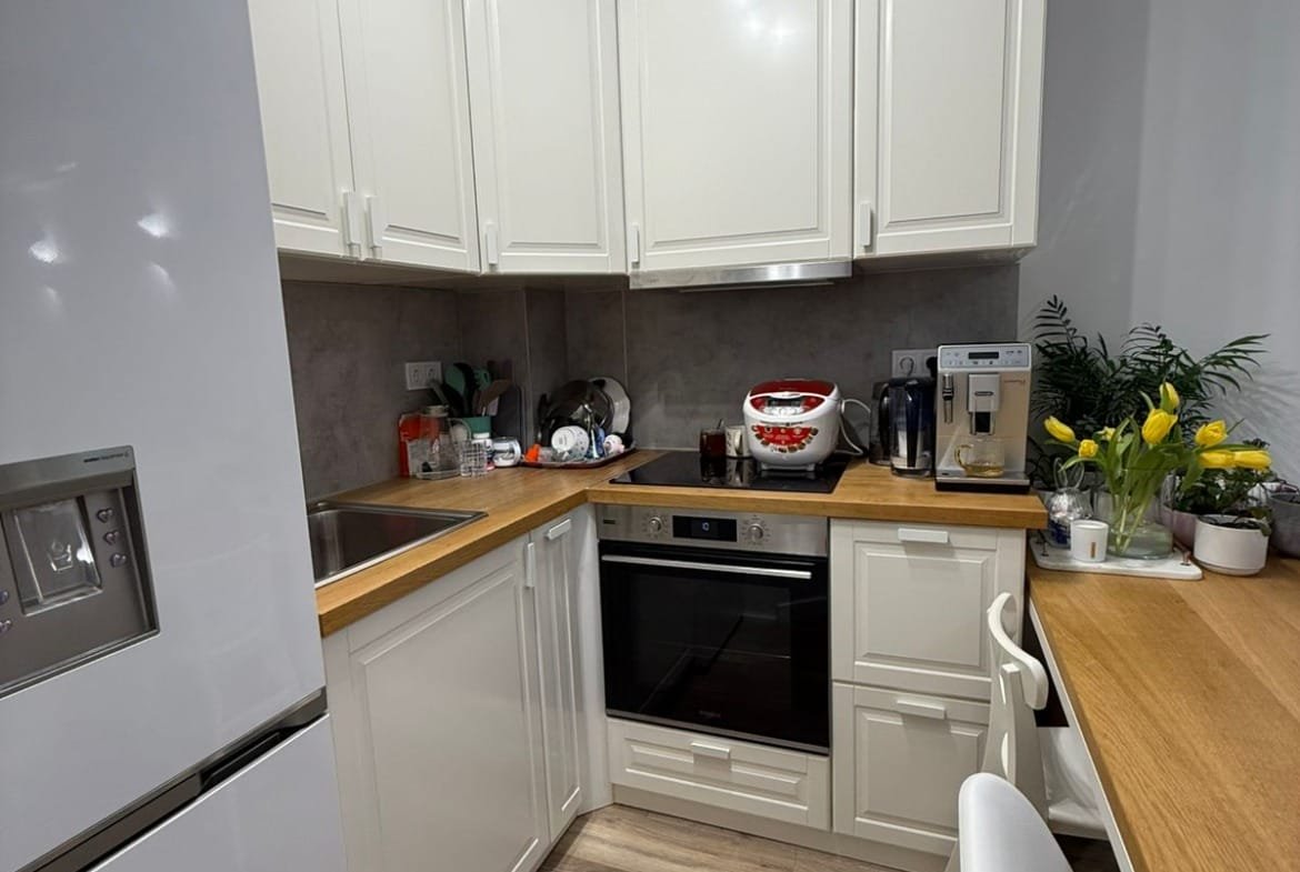 Современная кухня в двухкомнатном апартаменте с белыми шкафами, деревянным столом и техникой, включая холодильник и плиту.