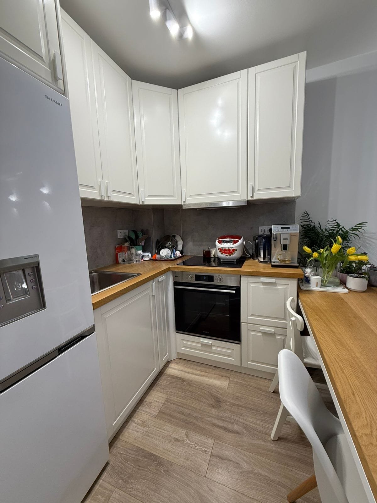 Современная кухня в двухкомнатном апартаменте с белыми шкафами, деревянным столом и техникой, включая холодильник и плиту.