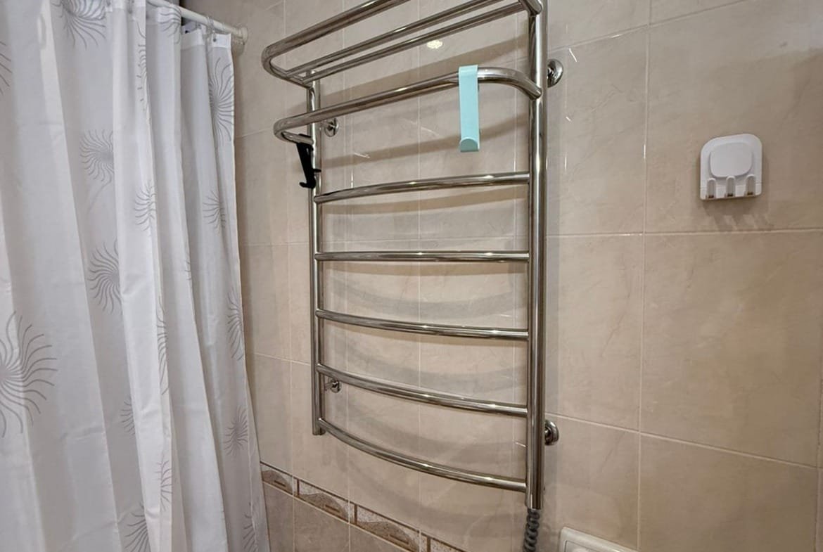 Стальная полотенцесушилка в ванной комнате с белой шторкой и плиткой светлых тонов.
