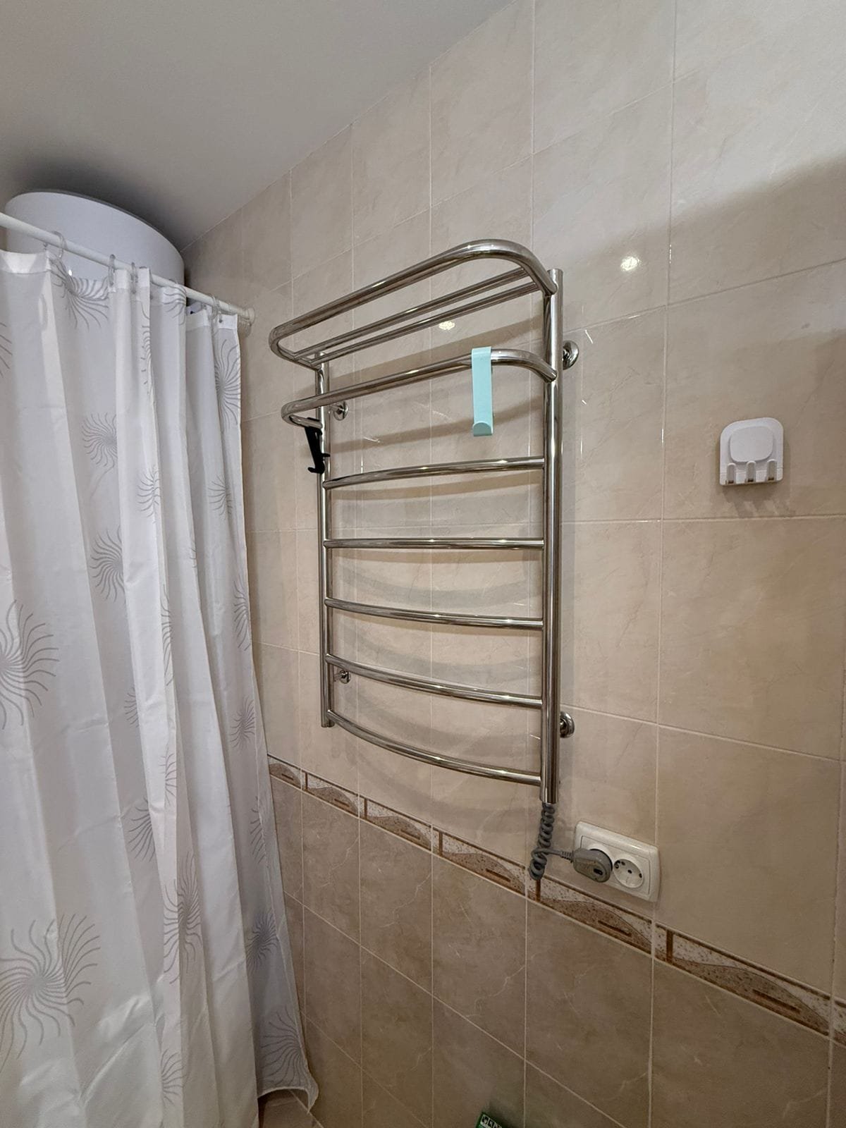 Стальная полотенцесушилка в ванной комнате с белой шторкой и плиткой светлых тонов.