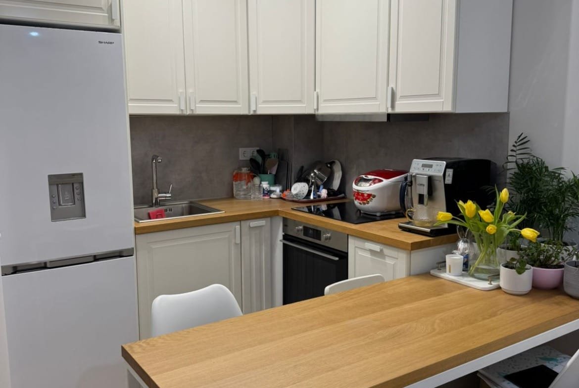 Современная кухня в двухкомнатном апартаменте с белыми шкафами, деревянным столом и зелеными растениями.