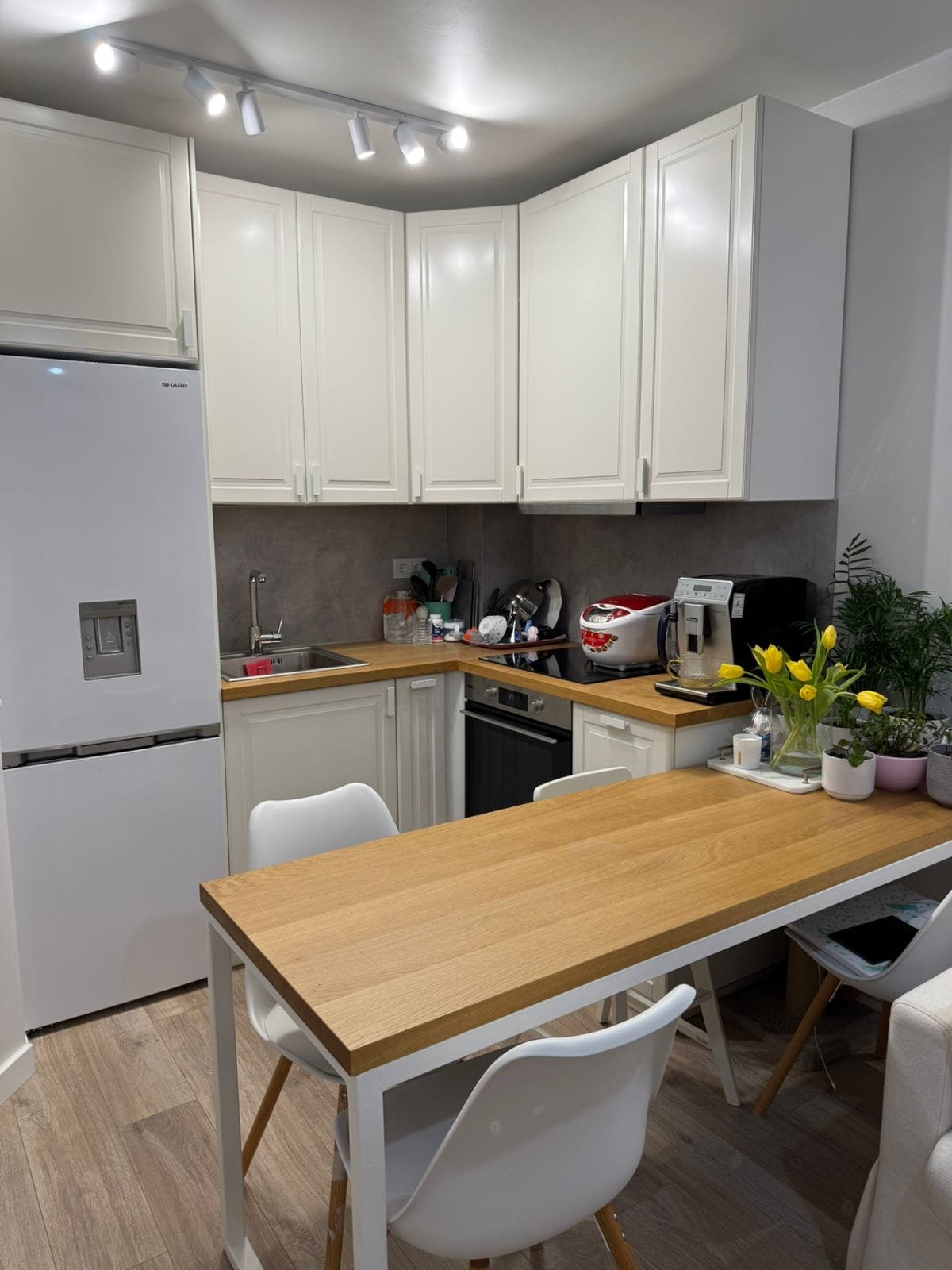 Современная кухня в двухкомнатном апартаменте с белыми шкафами, деревянным столом и зелеными растениями.
