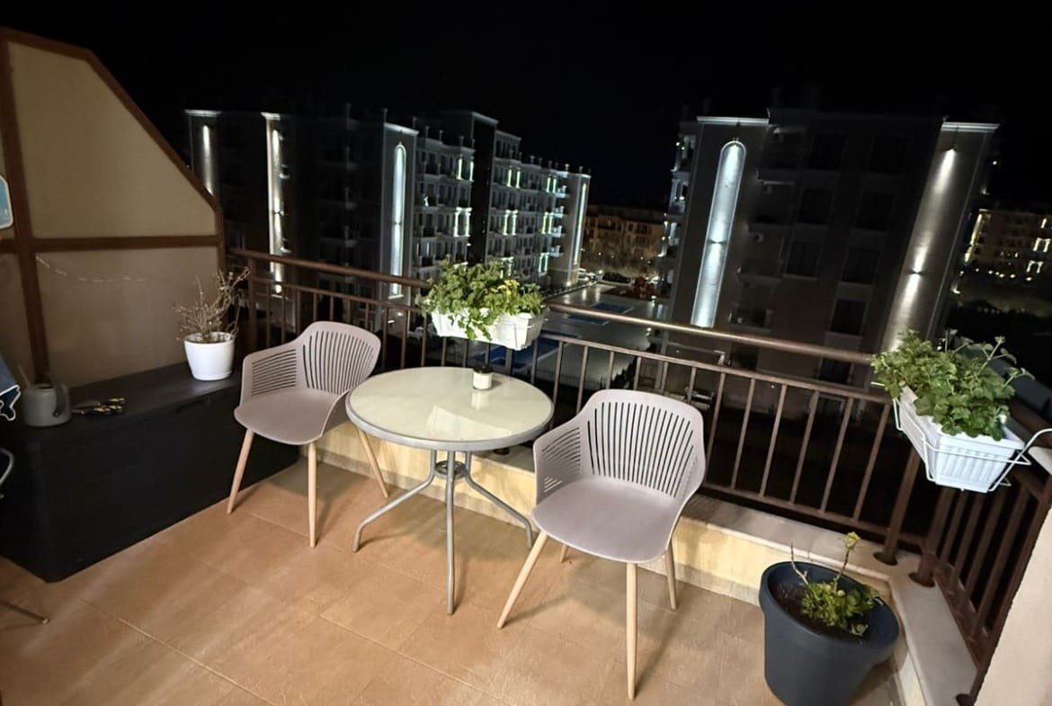 Балкон с двумя креслами и круглым столиком, зелеными растениями, ночной городской пейзаж на фоне.