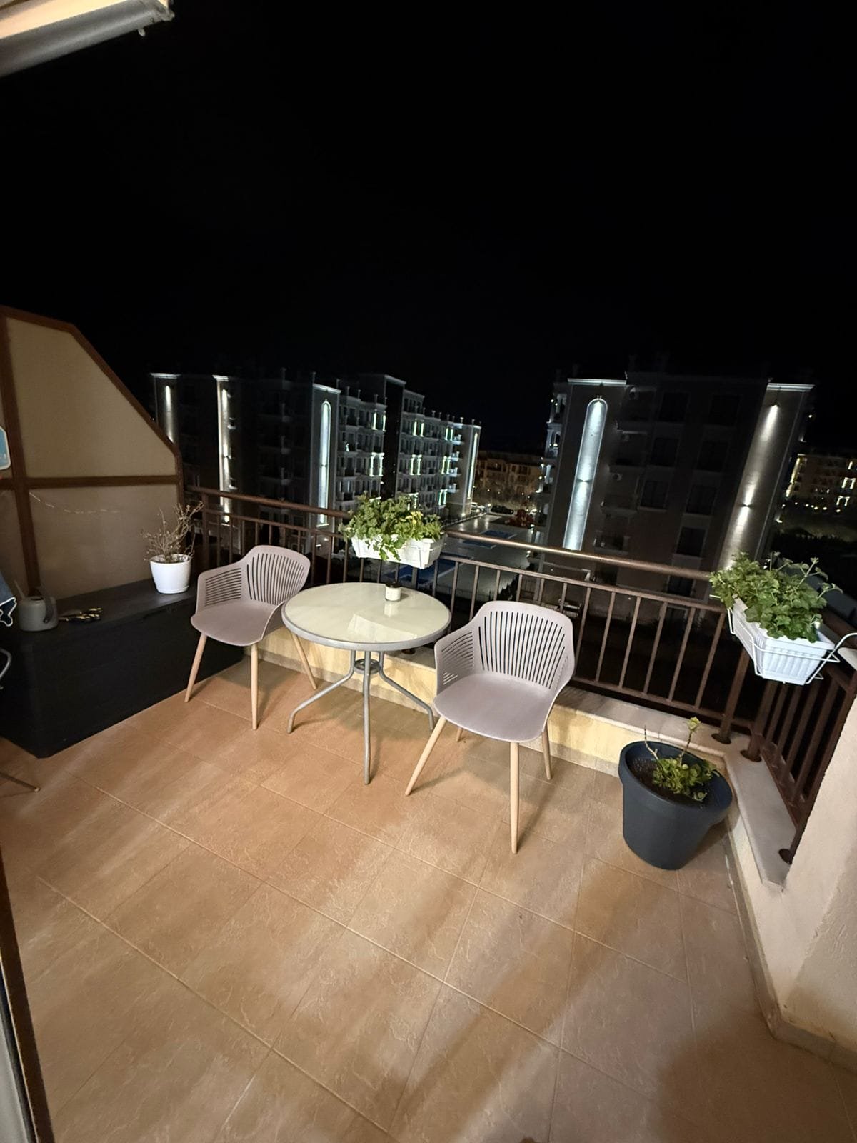Балкон с двумя креслами и круглым столиком, зелеными растениями, ночной городской пейзаж на фоне.