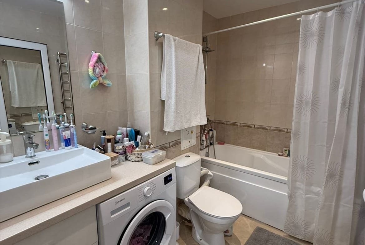 Современная ванная комната с ванной, туалетом, стиральной машиной и зеркалом, оформленная в светлых тонах.