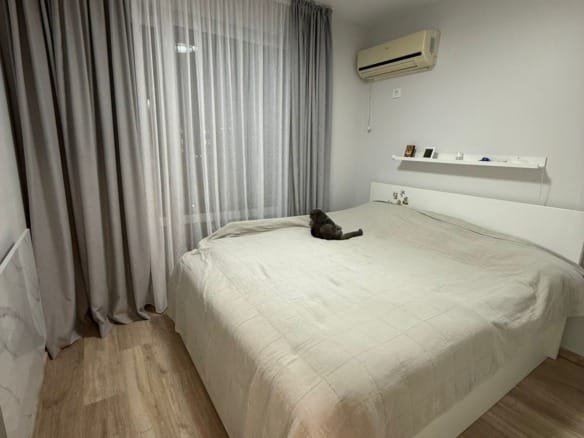 Современная спальня с двуспальной кроватью, светлыми шторами, паркетным полом и роботом-пылесосом на полу.