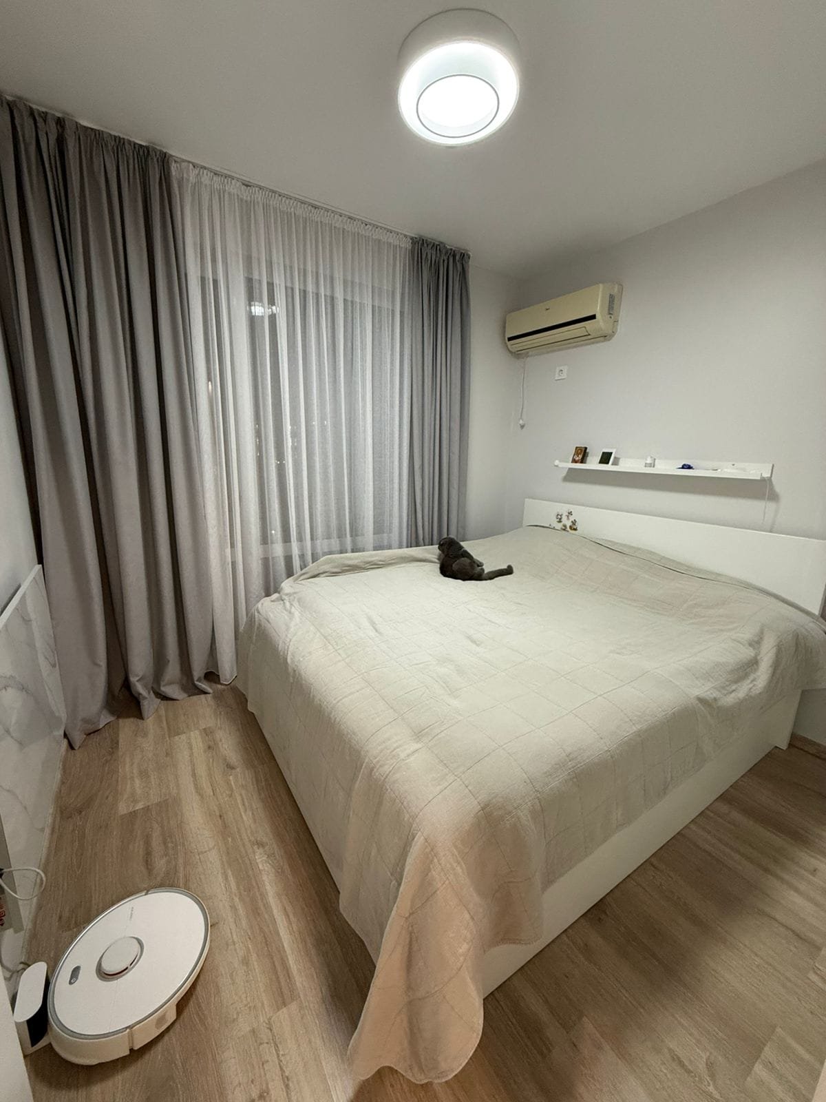 Современная спальня с двуспальной кроватью, светлыми шторами, паркетным полом и роботом-пылесосом на полу.