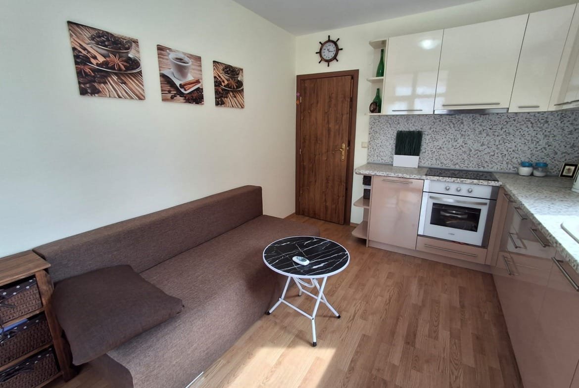 Современный интерьер квартиры с угловым диваном, журнальным столиком и кухонной зоной с плитой и темной столешницей.