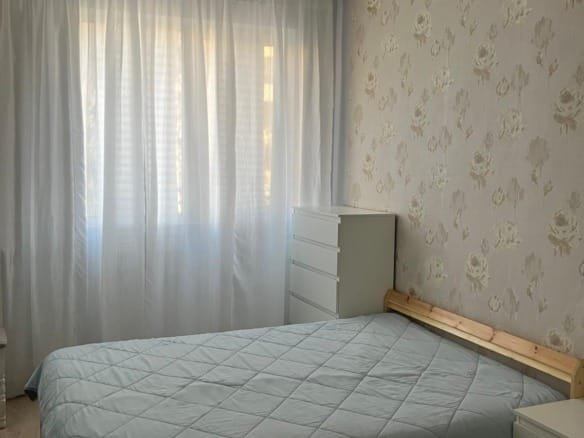 Светлая спальня с большим окном, белыми шторами, двуспальной кроватью и комодом, подходит для комфортного проживания в Болгарии.