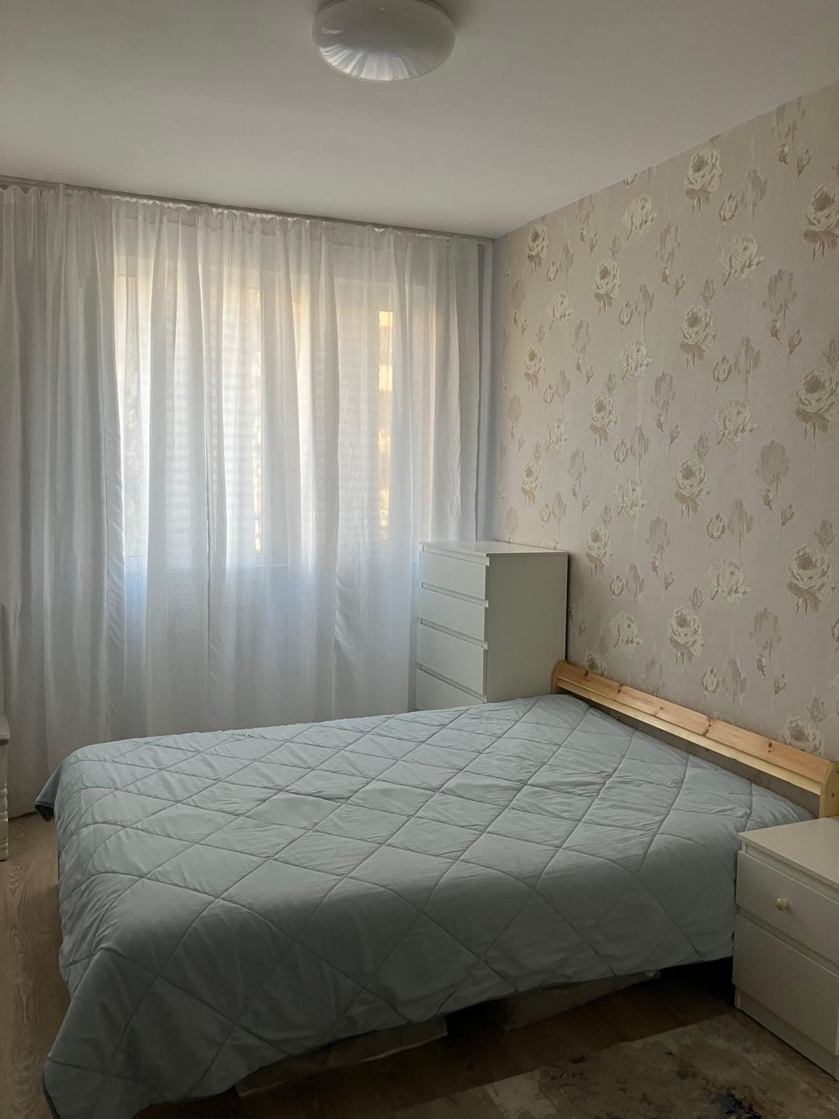 Светлая спальня с большим окном, белыми шторами, двуспальной кроватью и комодом, подходит для комфортного проживания в Болгарии.