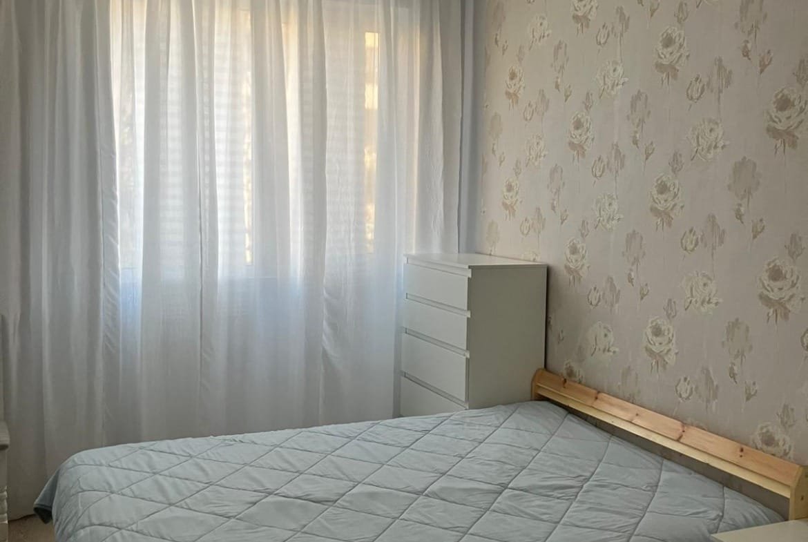 Уютная спальня с двуспальной кроватью, светлыми занавесками и светло-кремовыми обоями, идеальна для покупки недвижимости в Болгарии.