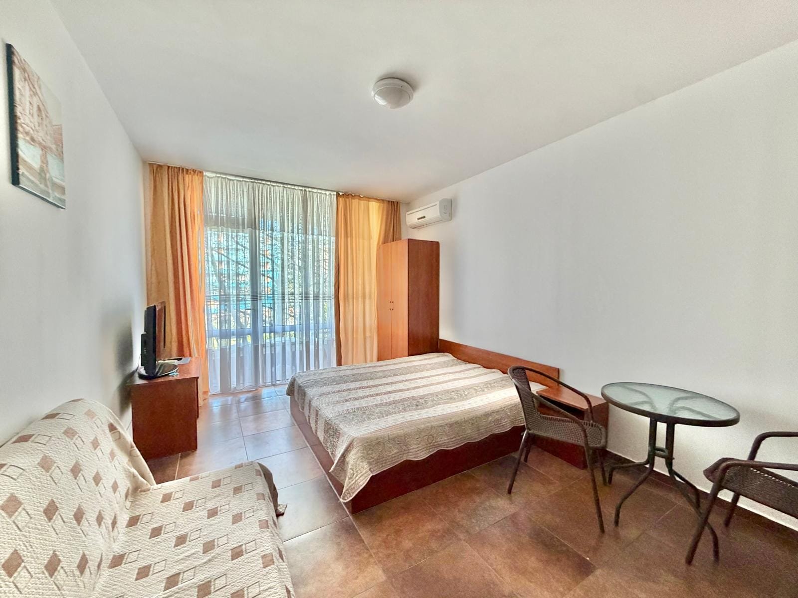 Современная квартира в Болгарии, светлая комната с двуспальной кроватью, телевизором и зоной отдыха у окна.