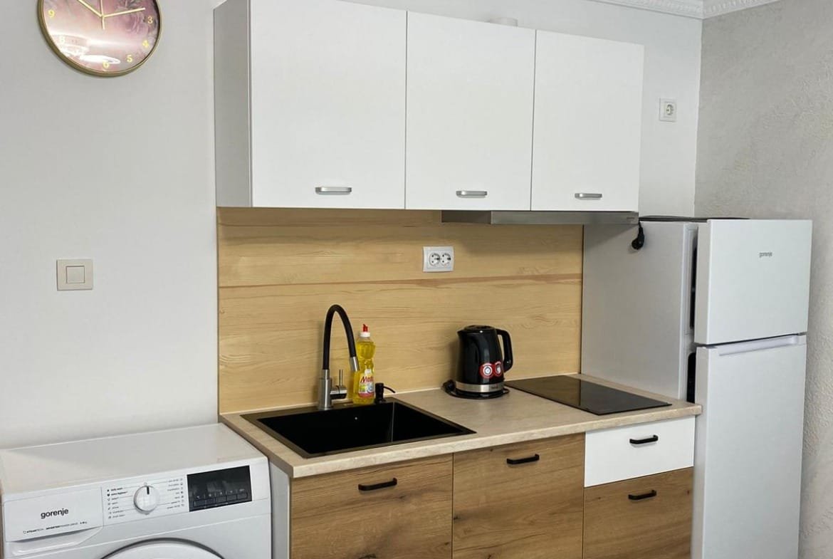 Современная кухня с гладкими белыми шкафами, стиральной машиной, холодильником и черной раковиной в интерьере Болгарской недвижимости.