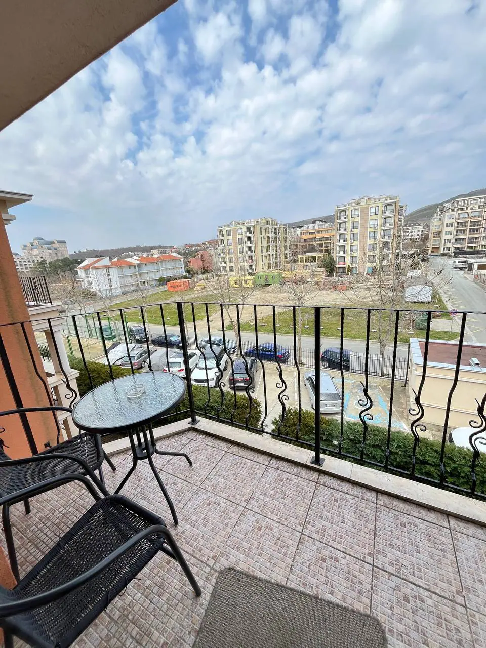 Балкон двухкомнатного апартамента с видом на комплекс “Prestige Fort Beach” Балкон с двумя черными стульями и круглым столиком, вид на зеленую зону и жилые здания комплекса Prestige Fort Beach.