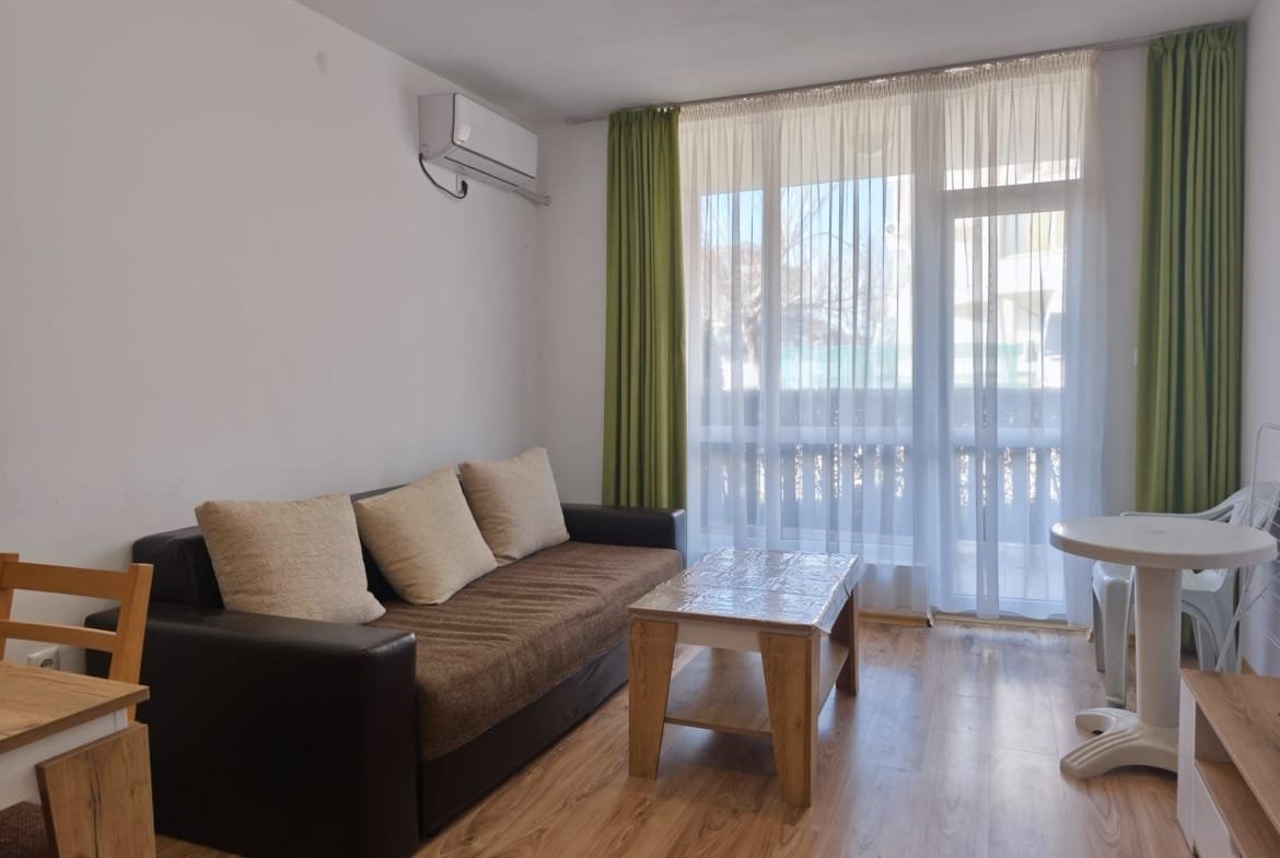 Современная гостиная в Болгарии с диваном, столом и большими окнами, создающими светлую и уютную атмосферу.